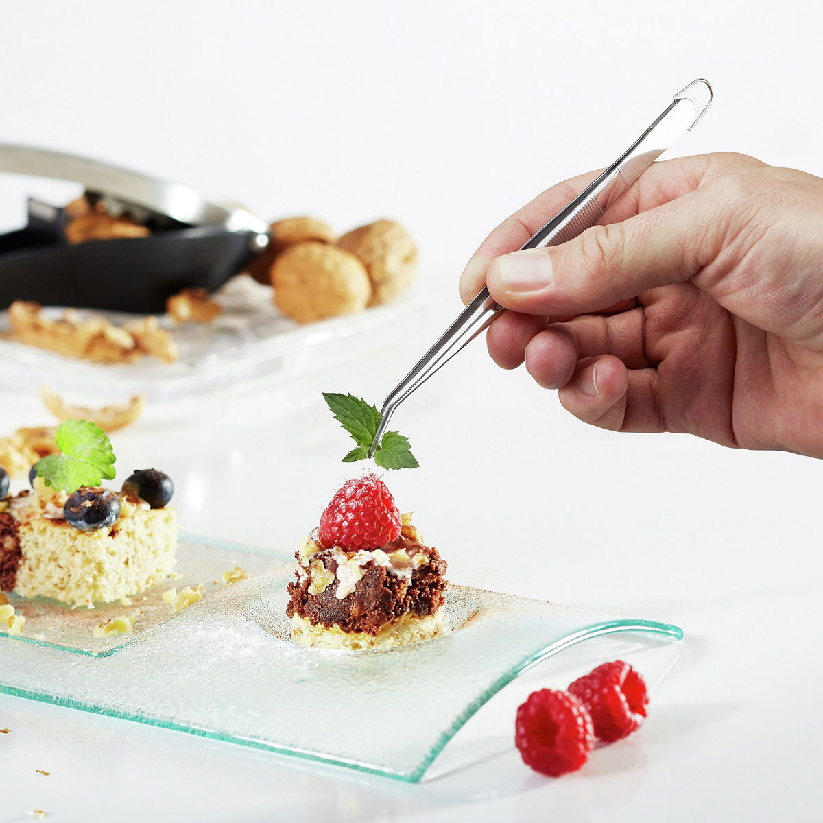Hand hält eine Küchenpinzette PRECISO, die ein Dessert mit Himbeere und Minzblatt dekoriert, seitliche Perspektive.
