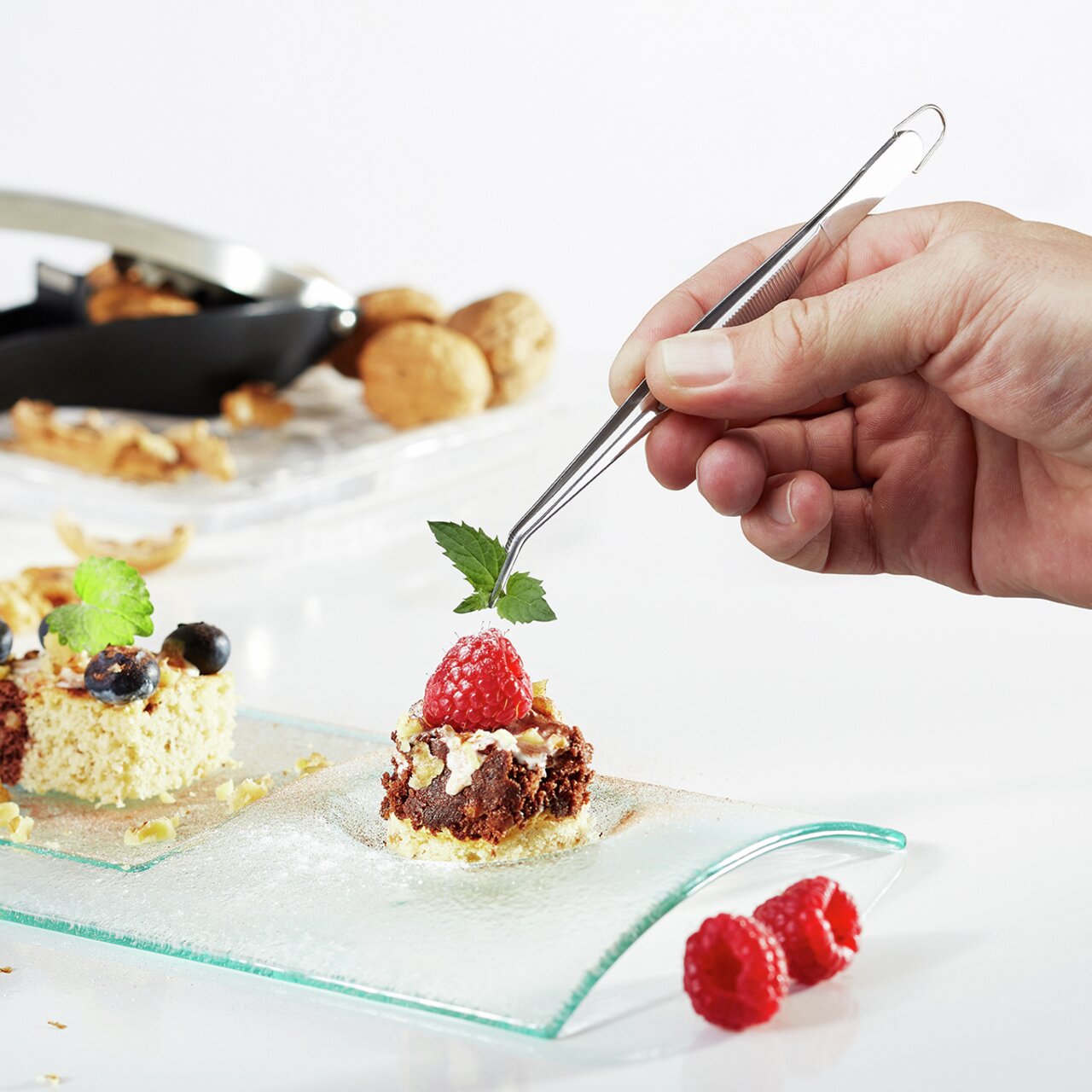 Hand hält eine Küchenpinzette PRECISO, die ein Dessert mit Himbeere und Minzblatt dekoriert, seitliche Perspektive.