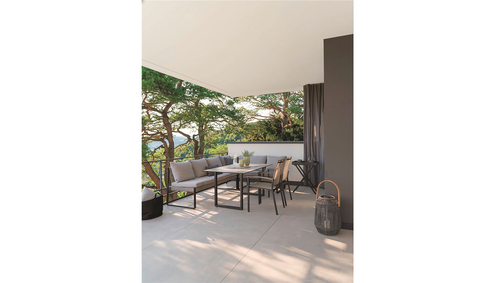 Stern Tuintafel KUFENTISCHSYSTEM Moderner Gartentisch mit Stühlen und Lounge-Sofa auf einer Terrasse mit Blick auf Bäume, aus seitlicher Perspektive fotografiert.