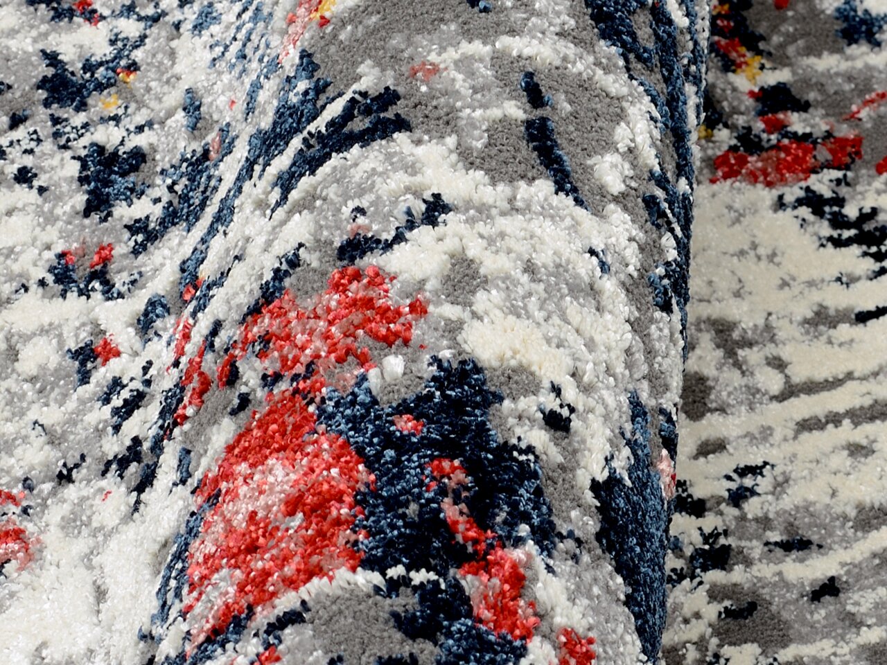 Nahaufnahme eines Designer-Teppichs mit abstraktem Muster in Grau, Blau, Rot und Weiß, aus seitlicher Perspektive aufgenommen.