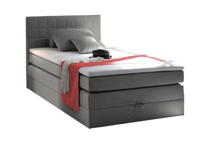 Graues Boxspringbett mit Kopfteil, seitliche Perspektive, dekoriert mit Kissen und rotem Überwurf.