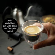 Nahaufnahme eines doppelwandigen Glases mit Espresso, gehalten in einer Hand, im Hintergrund eine Kaffeekanne. Kein Verbrühen am Glas dank der Doppelwand.