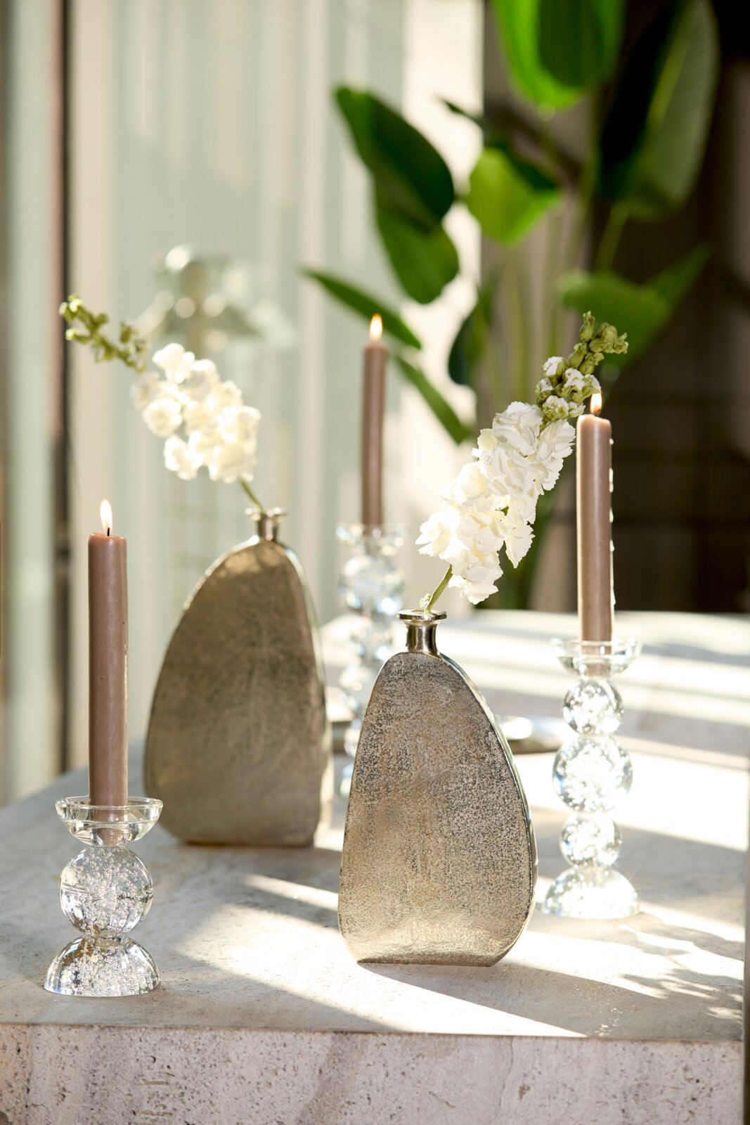 Vooraanzicht van een decoratieve zilverkleurige vaas met witte bloemen, stijlvol opgesteld naast brandende taupekleurige kaarsen in glazen kandelaars voor een spiegel.