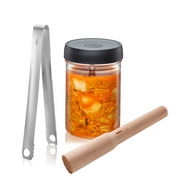 Fermentierset Nativo bestehend aus einem Glas mit schwarzem Deckel, gefüllt mit fermentiertem Gemüse, einer Edelstahlzange und einem hölzernen Stampfer, aus der Frontalperspektive.