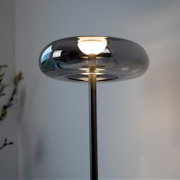 Moderne Stehlampe mit rauchgrauem, ovalem Glasschirm und warm leuchtender Lichtquelle auf schlankem Metallfuß, fotografiert in Frontansicht aus leicht niedriger Perspektive