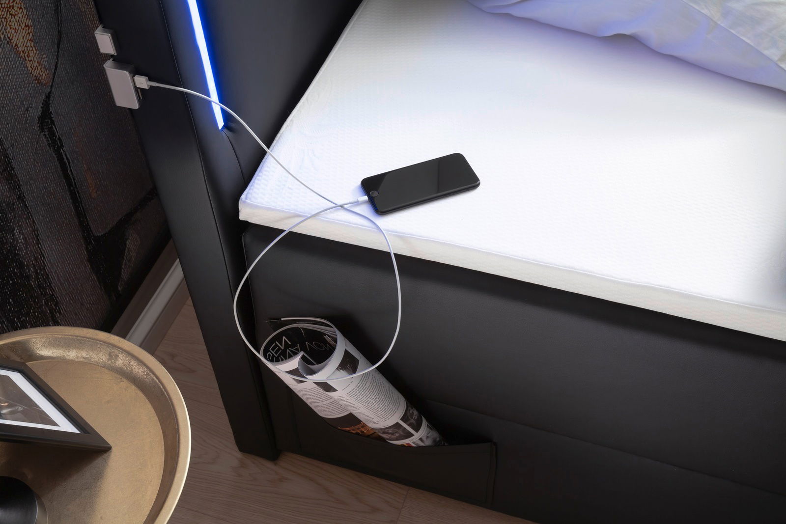 Nahaufnahme eines Boxspringbetts mit integrierter LED-Beleuchtung, USB-Ladeanschluss und seitlicher Aufbewahrungstasche, aus der eine Zeitschrift ragt. Perspektive von oben.