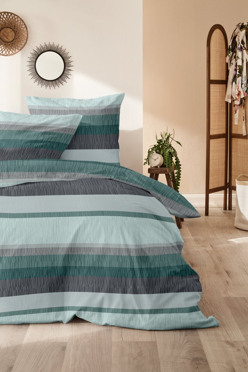 Bettwäsche-Set in Blau- und Grautönen mit Streifenmuster, auf einem Bett in einem gemütlichen Schlafzimmer mit Holzboden und Dekorationen, aus seitlicher Perspektive fotografiert.