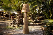 Akku-Outdoor-Stehleuchte aus Rattan in einem Garten mit Palmen, seitliche Perspektive