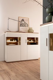 Modernes weißes Sideboard mit Holzplatte und integrierter LED-Beleuchtung in den offenen Fächern, dekoriert mit Tischlampe, gerahmtem Poster und Glasvase, fotografiert in leicht schräger Frontansicht aus Augenhöhe.