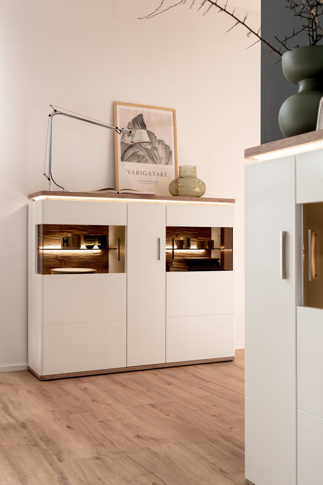 Modernes weißes Sideboard mit Holzplatte und integrierter LED-Beleuchtung in den offenen Fächern, dekoriert mit Tischlampe, gerahmtem Poster und Glasvase, fotografiert in leicht schräger Frontansicht aus Augenhöhe.