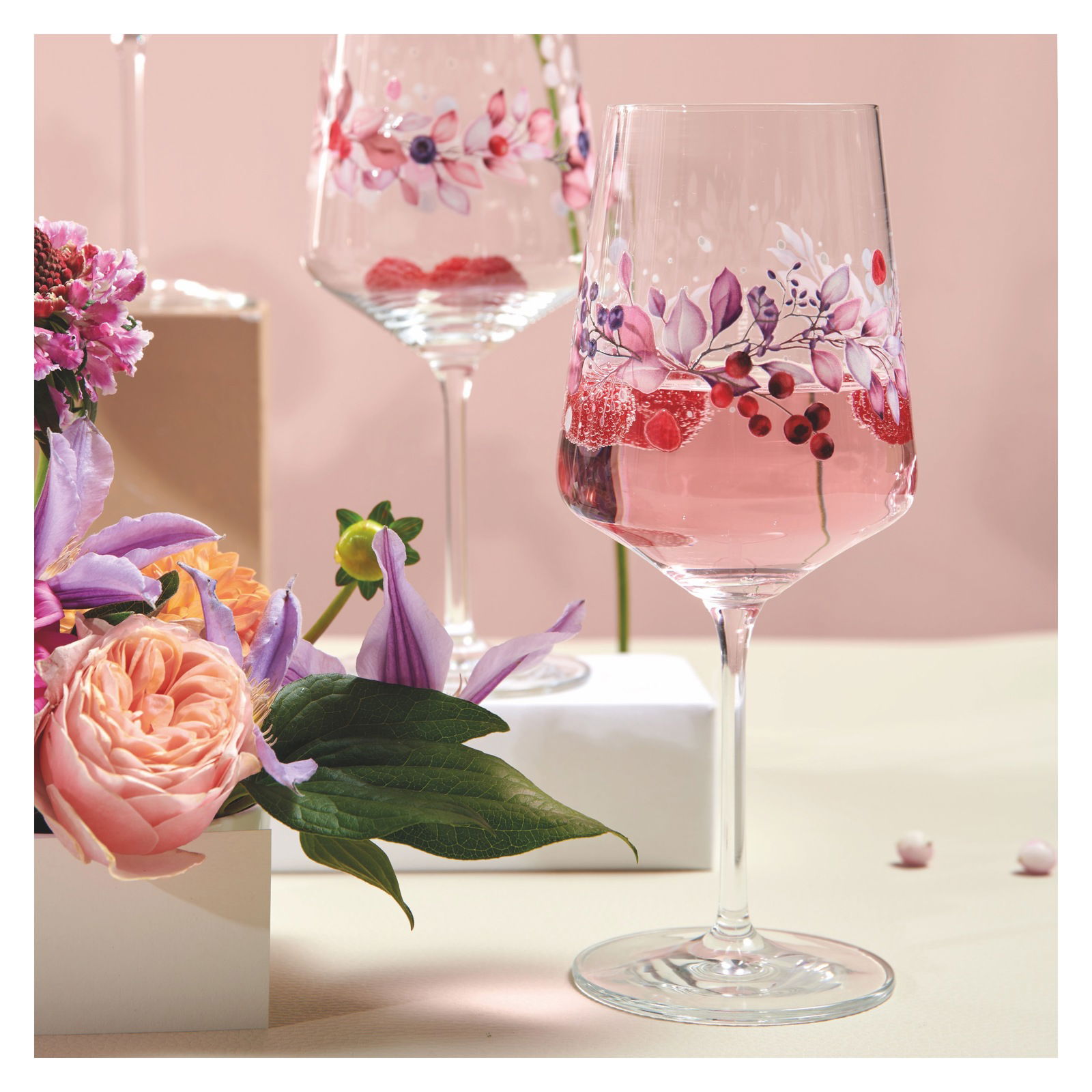Ein Weinglas mit floralen Mustern und rosa Getränk, umgeben von Blumen, aus einer seitlichen Perspektive fotografiert.