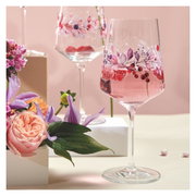 Ein Weinglas mit floralen Mustern und rosa Getränk, umgeben von Blumen, aus einer seitlichen Perspektive fotografiert.