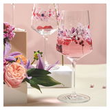 Ein Weinglas mit floralen Mustern und rosa Getränk, umgeben von Blumen, aus einer seitlichen Perspektive fotografiert.