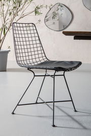 Moderner schwarzer Drahtstuhl mit Sitzpolster, seitliche Perspektive, vor einer hellen Wand mit Pflanzen und Spiegeln.