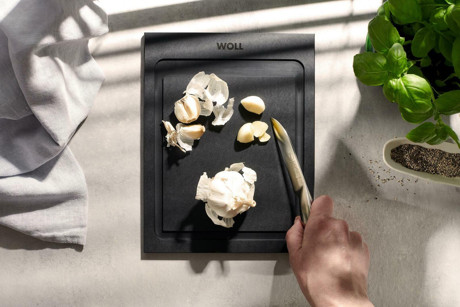 Schwarzes Schneidebrett mit Knoblauch und Messer, von oben fotografiert, neben Basilikum und Pfeffer.