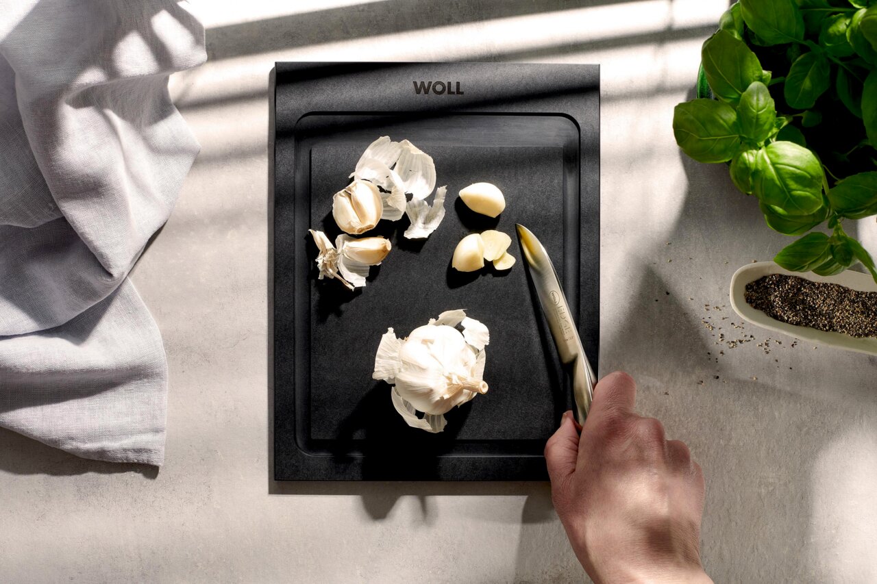 Woll Schneidebrett CULT & SERVE Schwarzes Schneidebrett mit Knoblauch und Messer, von oben fotografiert, neben Basilikum und Pfeffer.