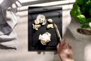 Schwarzes Schneidebrett mit Knoblauch und Messer, von oben fotografiert, neben Basilikum und Pfeffer.