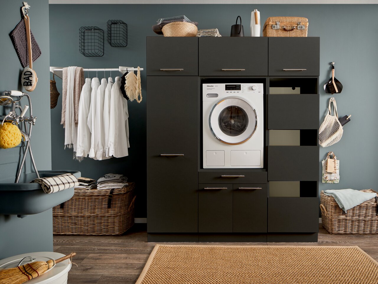 XORA Aufsatzschrank schmal PRIMUS Moderne Waschküche mit dunklem Hochschrank, integriertem Waschgerät und offenen Schubladen, frontal betrachtet.