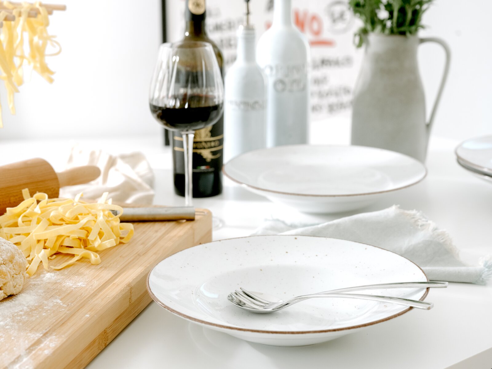 Schlichte weiße, gesprenkelte Pastaschale mit braunem Rand und zwei Löffeln auf weißem Tisch, im Vordergrund aus leicht schräger Seitenansicht; im Hintergrund Nudelbrett mit Bandnudeln, Rotweinglas, Flaschen und Vase.