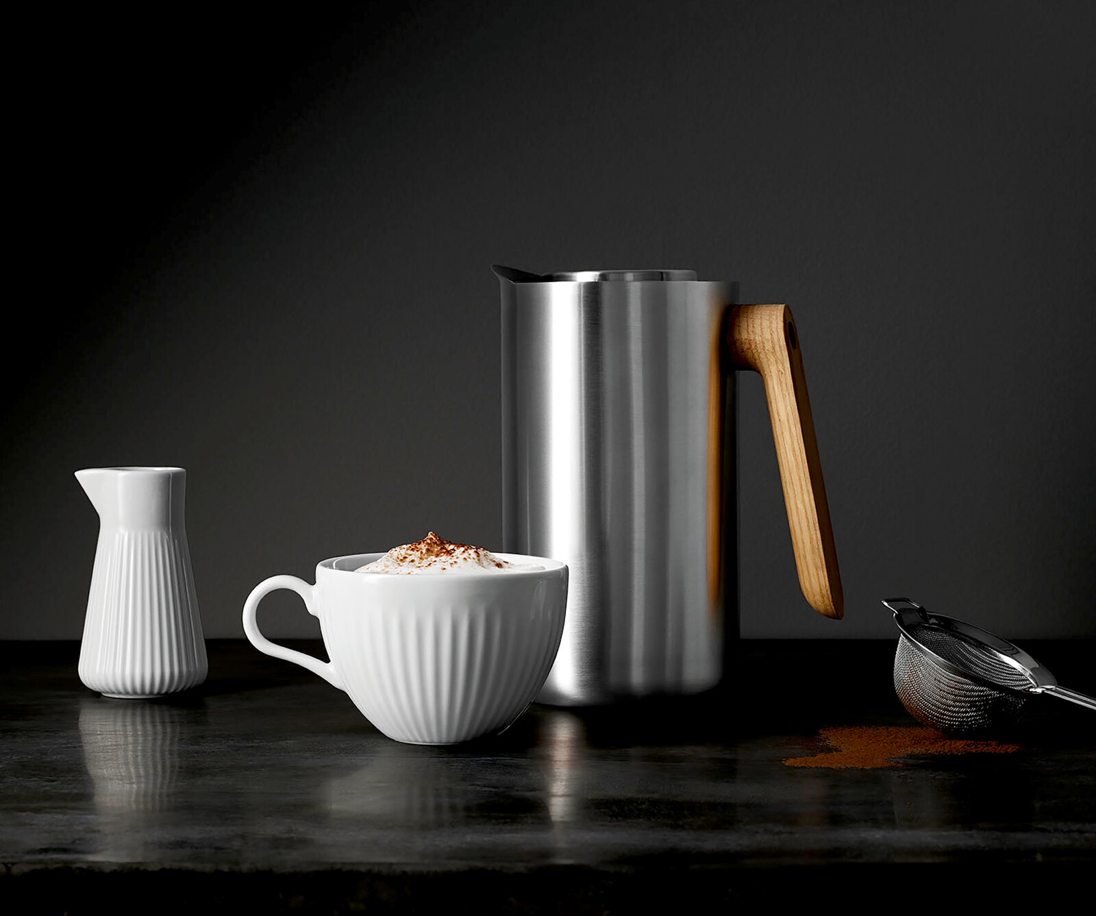 Eva Solo Isolierkanne NORDIC KITCHEN Edelstahl-Milchaufschäumkännchen mit Holzgriff neben weißer geriffelter Tasse mit Milchschaum und Kakaopulver, kleiner weißer Milchkanne und Sieb auf dunkler Oberfläche, Frontansicht auf Augenhöhe