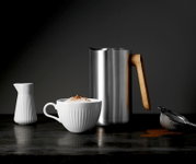 Roestvrijstalen melkopschuimkan met houten handvat op donker aanrecht, met witte kop cappuccino, wit melkkannetje en fijne zeef met cacaopoeder; zijaanzicht op tafelhoogte