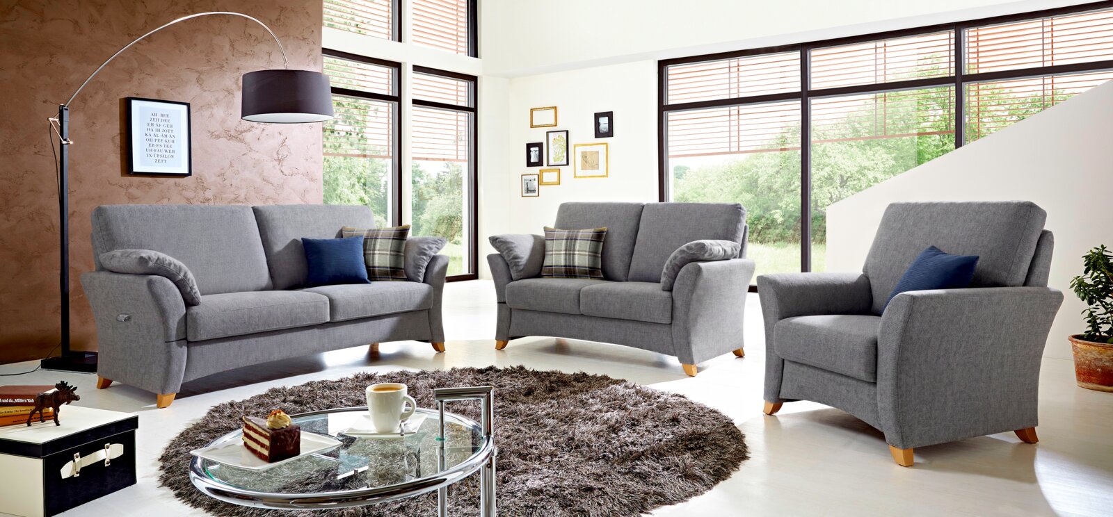 Graue Polstergarnitur im Wohnzimmer: 3-Sitzer-Sofa, 2-Sitzer-Sofa und Sessel mit Stoffbezug und Holzfüßen, frontal in leichter Schrägansicht fotografiert.