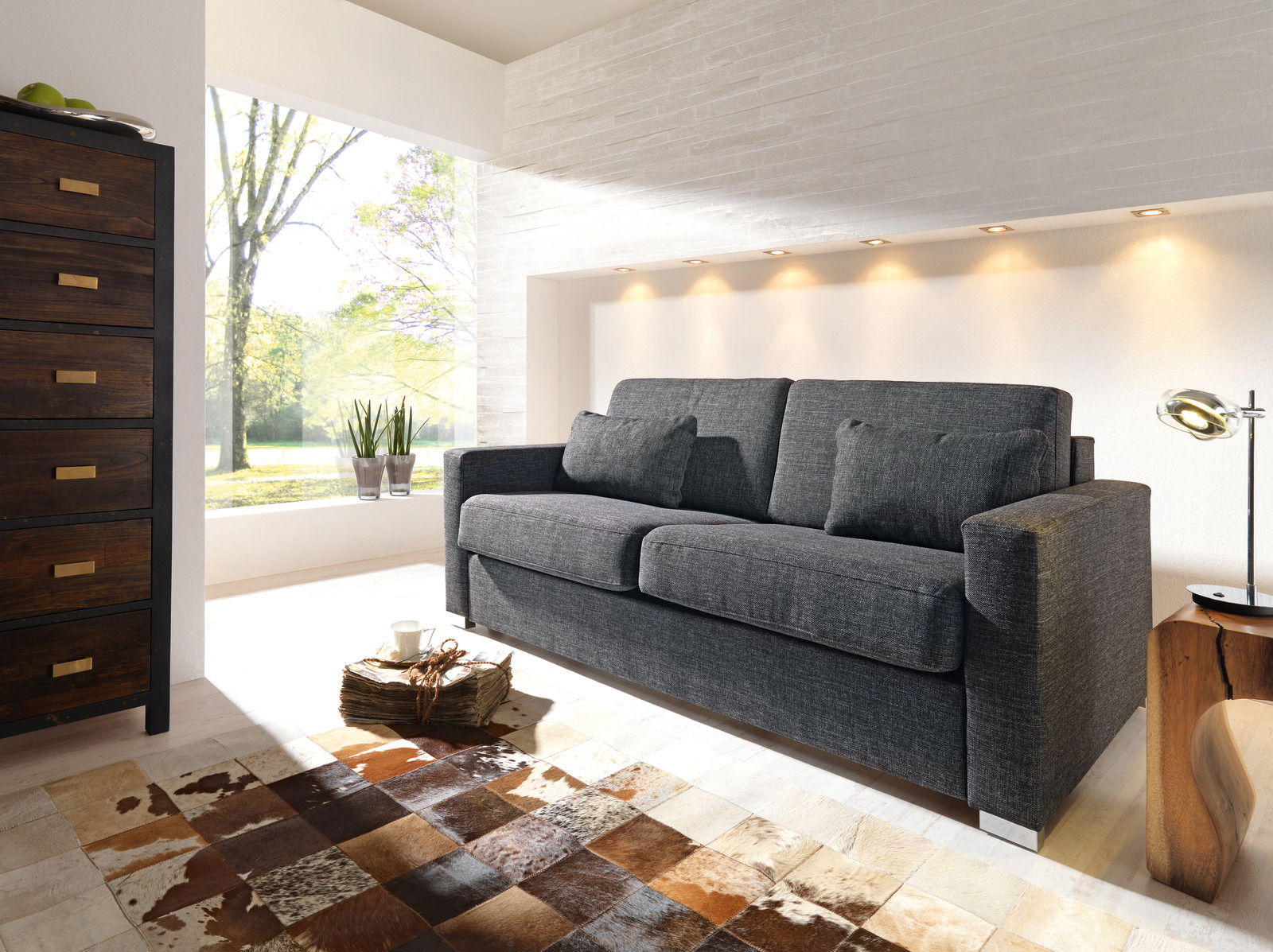 Novel Schlafsofa BRUTUS Graues Verwandlungssofa in modernem Wohnzimmer, seitliche Perspektive, mit hellem Tageslicht und stilvoller Dekoration.