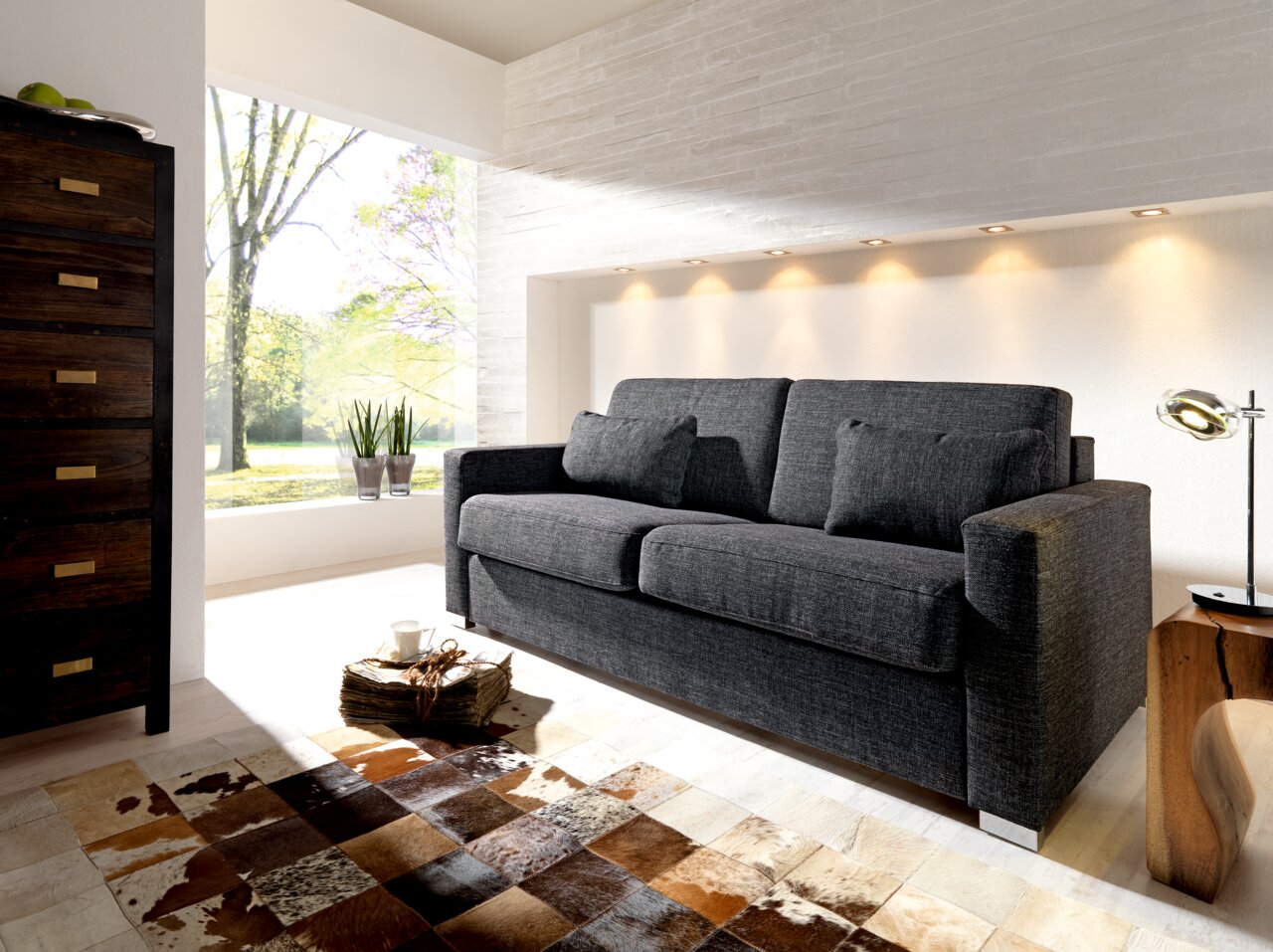 Novel Schlafsofa BRUTUS Graues Verwandlungssofa in modernem Wohnzimmer, seitliche Perspektive, mit hellem Tageslicht und stilvoller Dekoration.