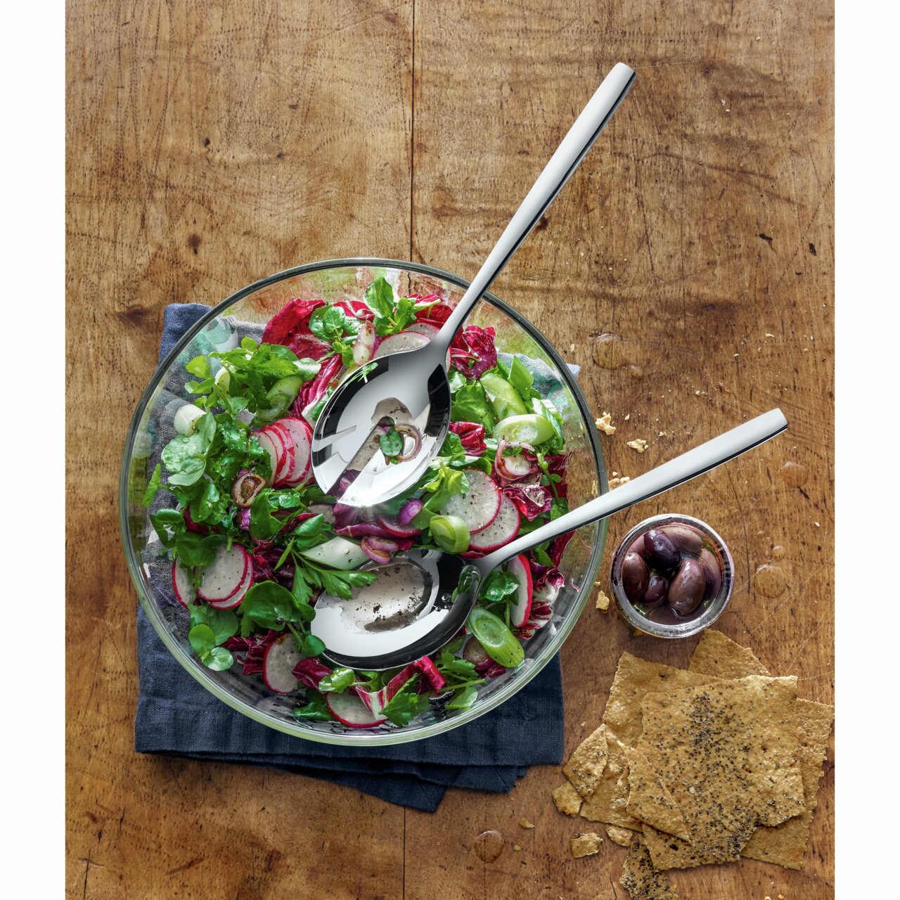 Vogelperspektive auf ein Salat-Set Taverno mit einer Glasschüssel voller frischem Salat, zwei glänzenden Salatbestecken und einer kleinen Schale mit Oliven auf einem Holztisch.