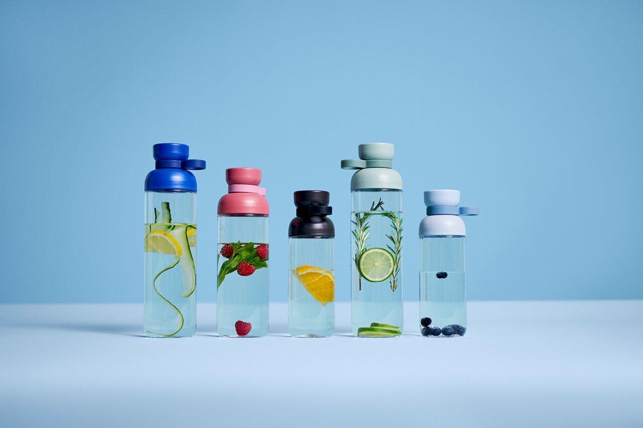 Fünf Trinkflaschen in verschiedenen Farben und mit verschiedenen Früchten gefüllt, auf einer blauen Oberfläche vor blauem Hintergrund, frontal betrachtet.