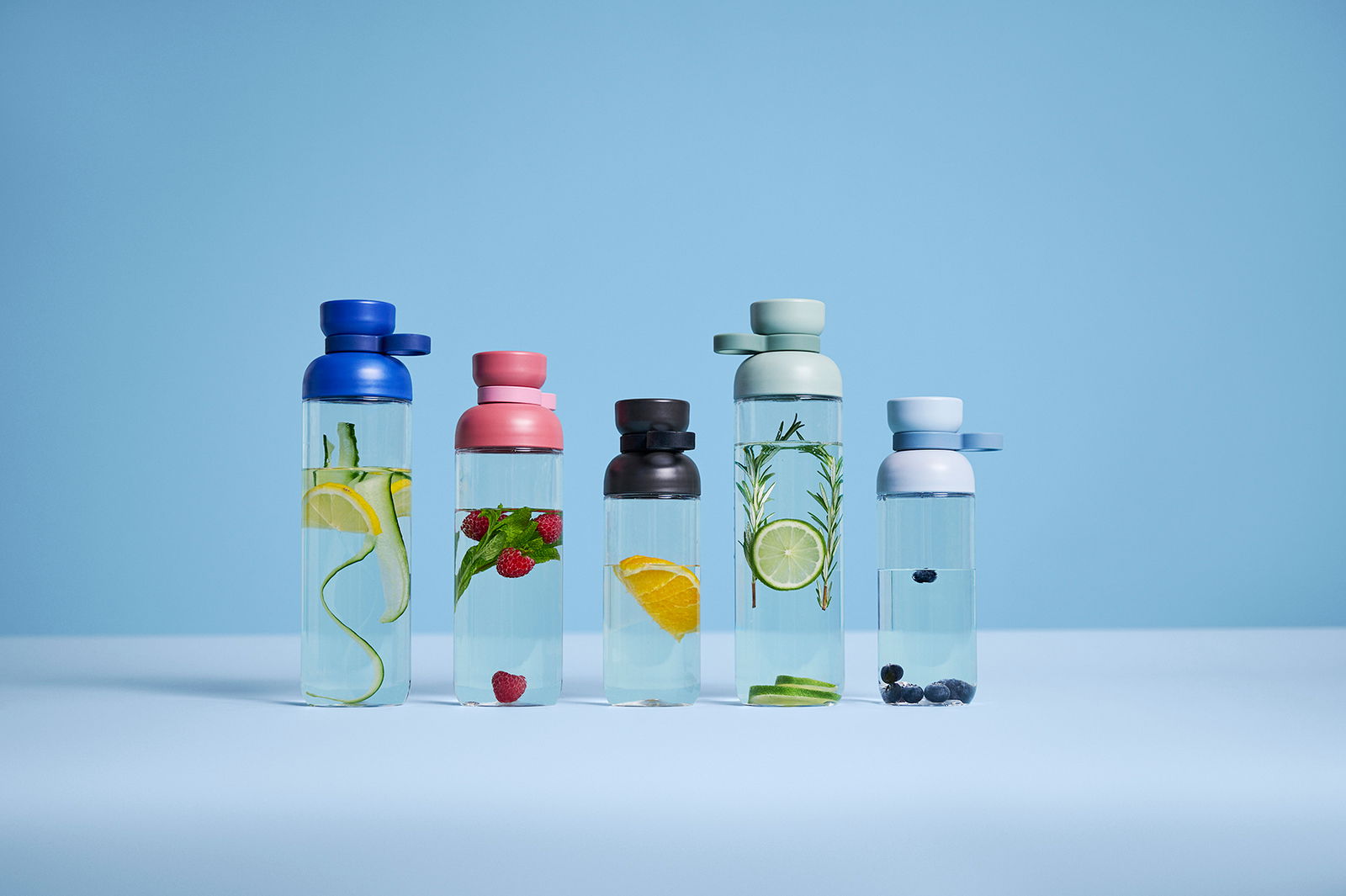 Fünf Trinkflaschen in verschiedenen Farben und mit verschiedenen Früchten gefüllt, auf einer blauen Oberfläche vor blauem Hintergrund, frontal betrachtet.