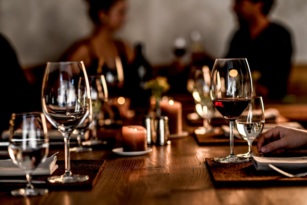 Elegante Bordeauxgläser auf einem festlich gedeckten Tisch, im Vordergrund ein Glas mit Rotwein, im Hintergrund unscharfe Personen bei einem Abendessen.