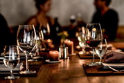 Elegante Bordeauxgläser auf einem festlich gedeckten Tisch, im Vordergrund ein Glas mit Rotwein, im Hintergrund unscharfe Personen bei einem Abendessen.