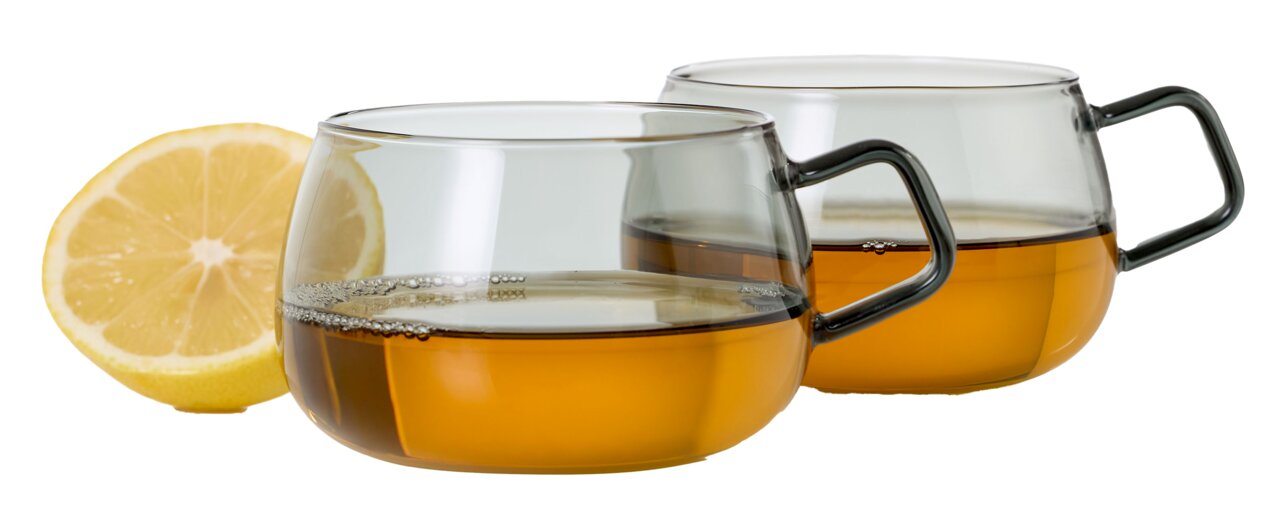 Zwei transparente Teetassen mit Henkel, gefüllt mit Tee, im Vordergrund eine halbe Zitrone. Seitliche Perspektive.