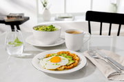Müslischale auf einem Tisch, gefüllt mit Salat, daneben ein Glas Wasser mit Minze, eine Tasse Kaffee und ein Teller mit Waffel, Spiegelei und Avocado. Perspektive von vorne.