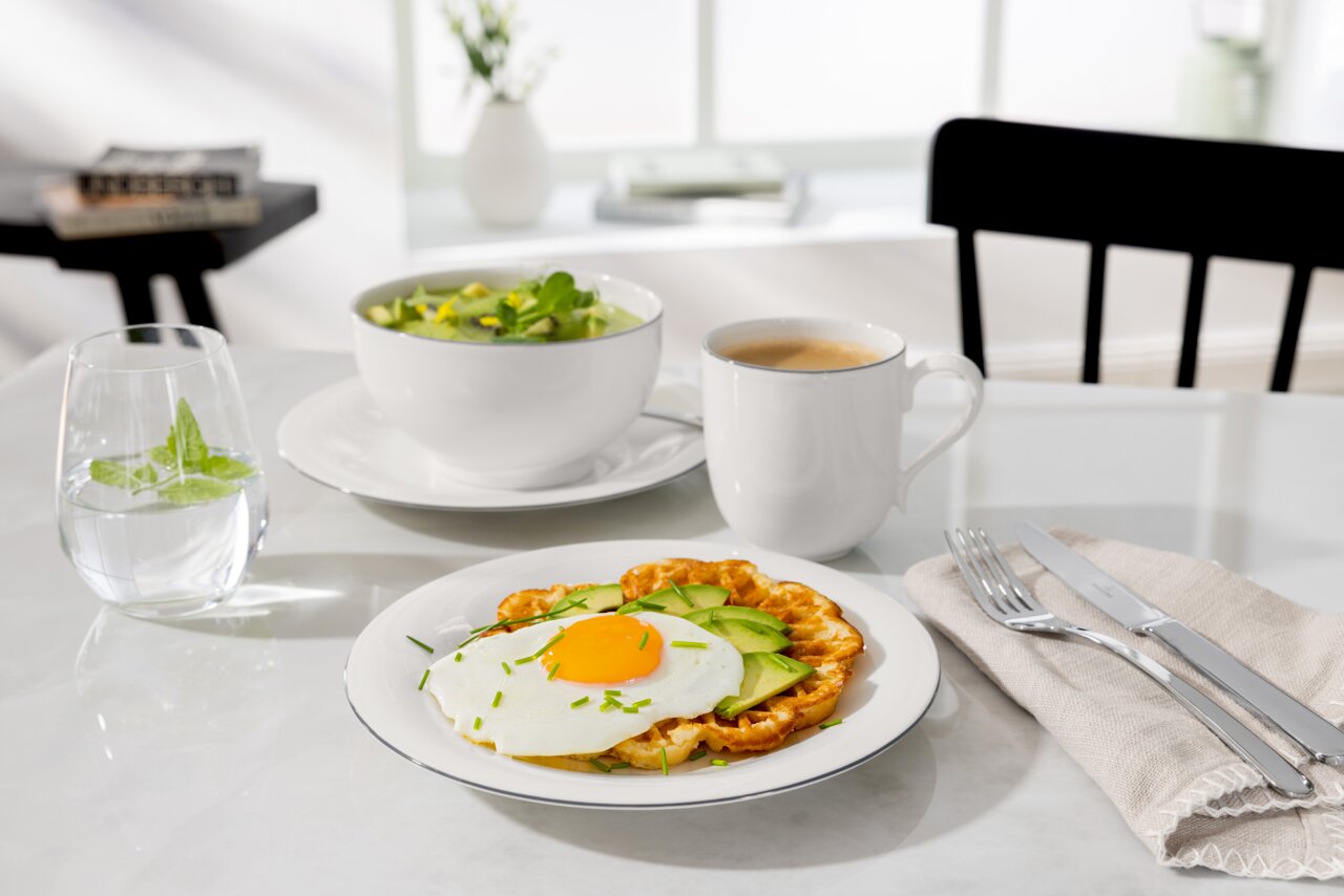 Müslischale auf einem Tisch, gefüllt mit Salat, daneben ein Glas Wasser mit Minze, eine Tasse Kaffee und ein Teller mit Waffel, Spiegelei und Avocado. Perspektive von vorne.