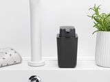Schwarzer Brabantia Seifenspender SINKSIDE auf einem weißen Waschbecken, frontal fotografiert, neben einem Handtuch und einer Pflanze.
