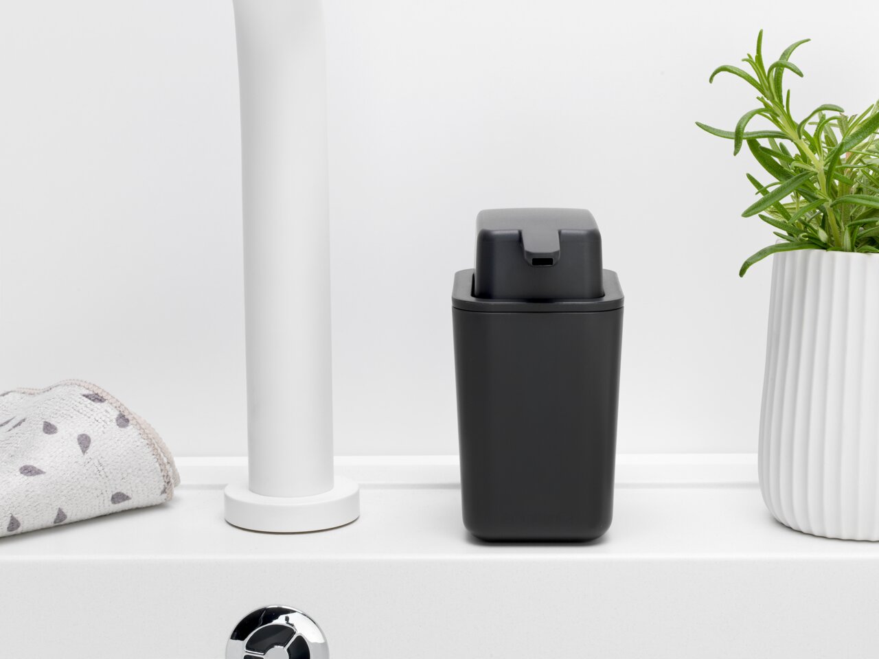 Een zwarte Brabantia-zeepdispenser SINKSIDE op een witte wastafel, van voren gefotografeerd, naast een handdoek en een plant.
