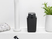 Een zwarte Brabantia-zeepdispenser SINKSIDE op een witte wastafel, van voren gefotografeerd, naast een handdoek en een plant.