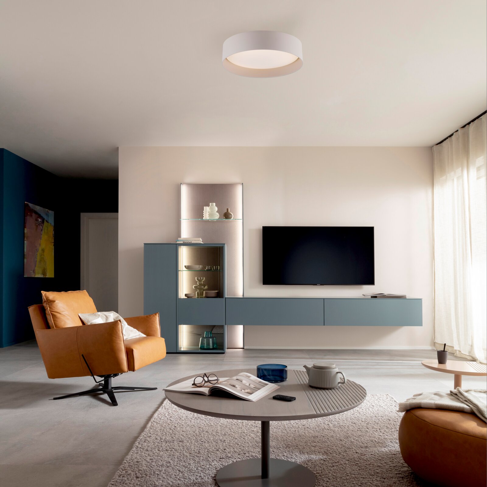 Modernes Wohnzimmer mit blauem TV-Lowboard und Vitrine, Wand-TV, cognacfarbenem Ledersessel und rundem Couchtisch; Ansicht aus Augenhöhe in leichter Weitwinkelperspektive