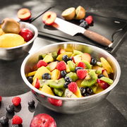 Edelstahlschüssel mit frischem Obstsalat, bestehend aus Kiwi, Himbeeren, Blaubeeren und Pfirsichen, auf einer Küchenarbeitsplatte. Im Hintergrund ein Schneidebrett mit Messer und weiteren Früchten. Perspektive von oben.