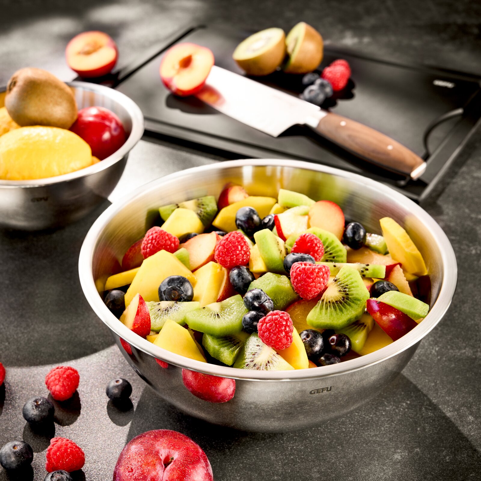 Edelstahlschüssel mit frischem Obstsalat, bestehend aus Kiwi, Himbeeren, Blaubeeren und Pfirsichen, auf einer Küchenarbeitsplatte. Im Hintergrund ein Schneidebrett mit Messer und weiteren Früchten. Perspektive von oben.