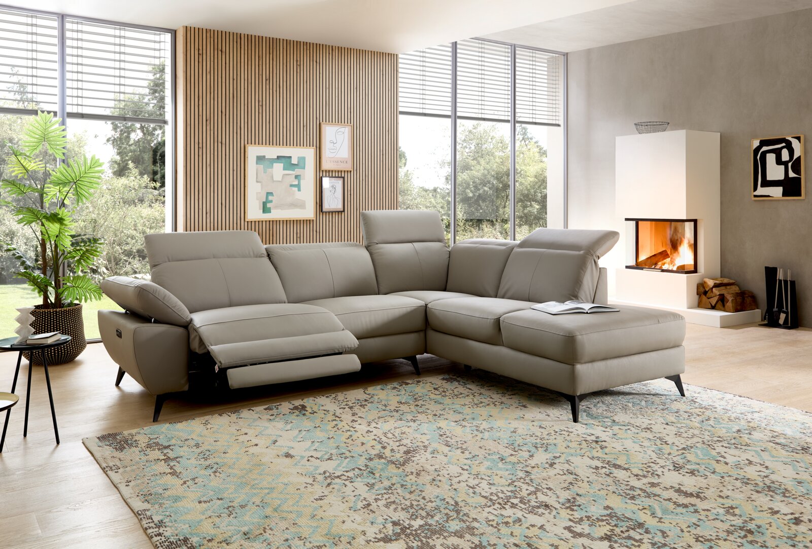 XORA Ecksofa PARIS Modernes graues Leder-Ecksofa mit Relaxfunktion und Ottomane im hellen Wohnzimmer mit großen Fenstern und Kamin, Perspektive: Frontansicht leicht schräg von links.