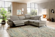 Modernes graues Leder-Ecksofa mit Relaxfunktion und Ottomane im hellen Wohnzimmer mit großen Fenstern und Kamin, Perspektive: Frontansicht leicht schräg von links.