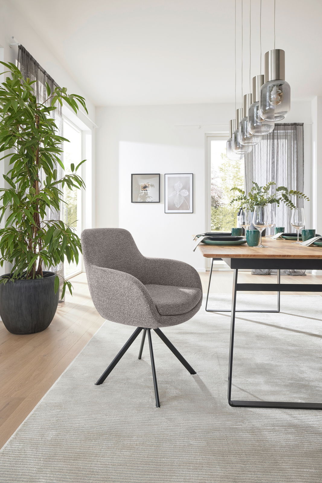 Moderner Armlehnstuhl mit grauem Stoffbezug und schwarzem Metallgestell, seitliche Perspektive in einem hellen Esszimmer mit Holztisch und Pflanzen.