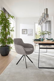Moderner Armlehnstuhl mit grauem Stoffbezug und schwarzem Metallgestell, seitliche Perspektive in einem hellen Esszimmer mit Holztisch und Pflanzen.