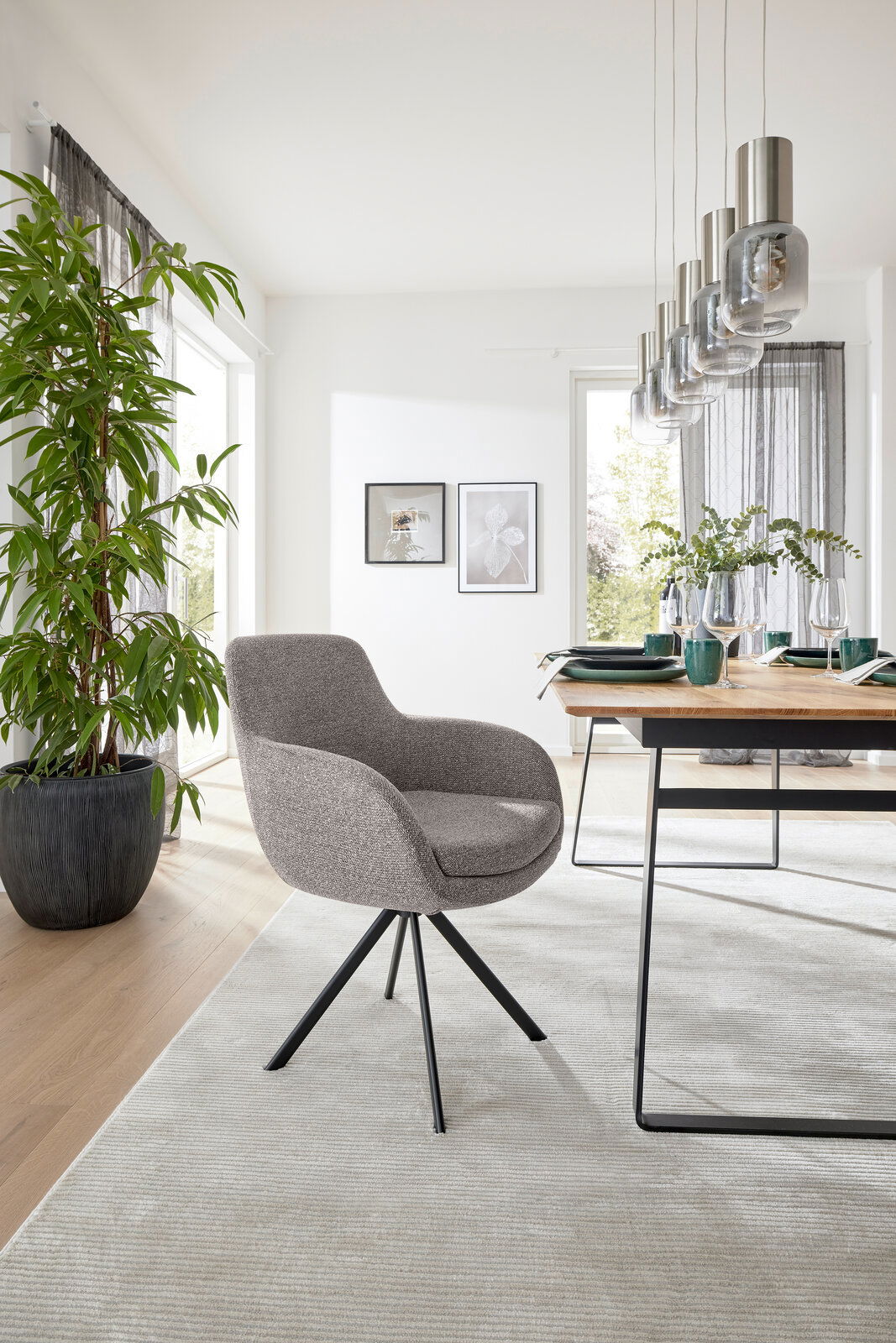 Moderner Armlehnstuhl mit grauem Stoffbezug und schwarzem Metallgestell, seitliche Perspektive in einem hellen Esszimmer mit Holztisch und Pflanzen.