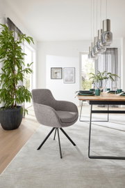Moderne fauteuil met grijze stoffen bekleding en een zwart metalen frame, zijaanzicht in een lichte eetkamer met een houten tafel en planten.