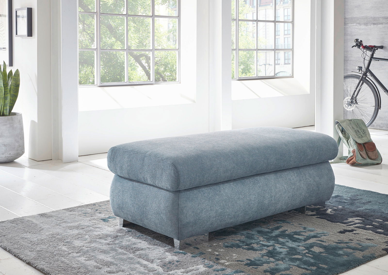 XXL-Hocker in hellblauem Stoff, seitliche Perspektive, in einem hellen Raum mit großen Fenstern und moderner Einrichtung.