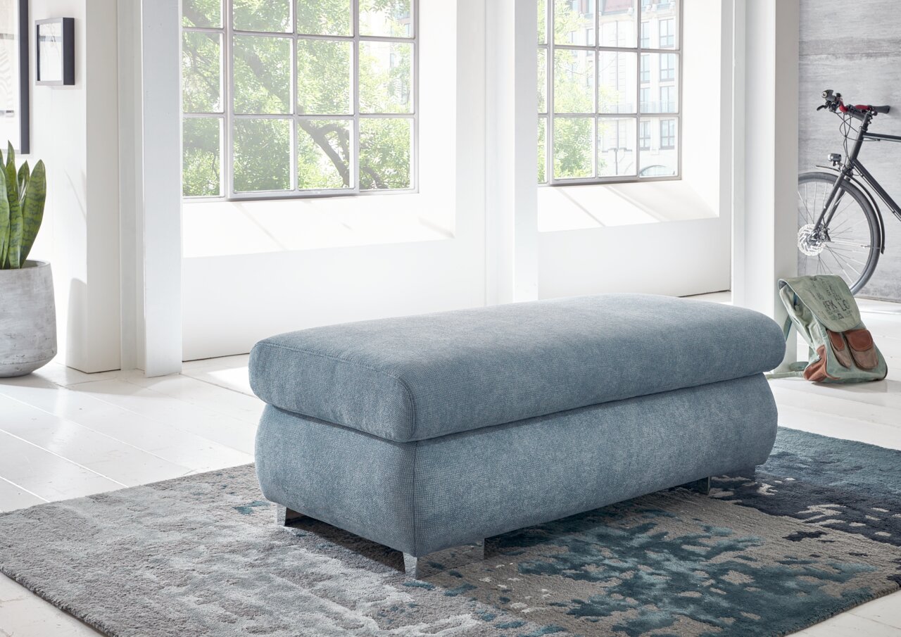 XXL-Hocker in hellblauem Stoff, seitliche Perspektive, in einem hellen Raum mit großen Fenstern und moderner Einrichtung.